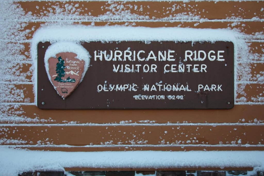 A wooden sign for the Hurricane Ridge Visitor Center in Olympic National Park, partially covered in snow, with the park's logo visible.