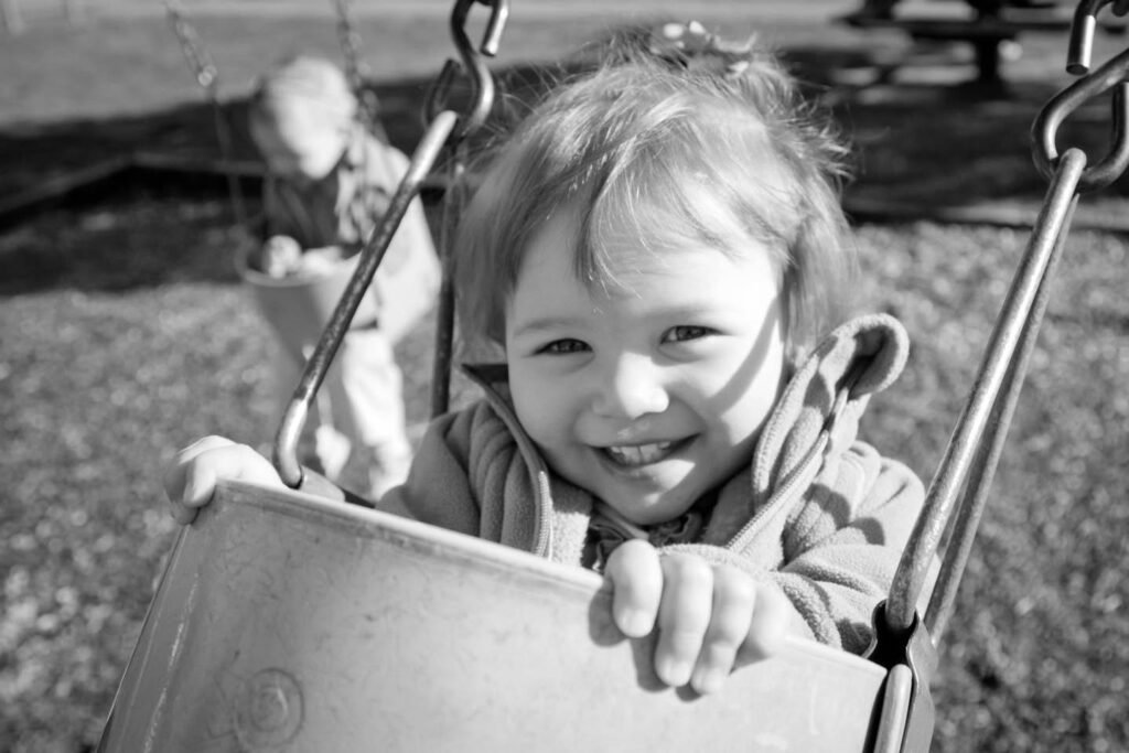 A smiling toddler in a bucket-style swing at a playground. A black and white photo capturing a moment of happiness.