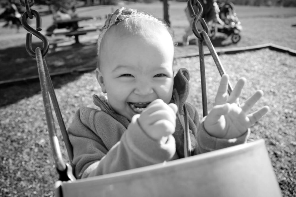 A happy child smiles and reaches from a playground bucket swing. Black and white close-up of metal chains and metal hooks attached to a swing.