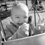 A happy child smiles and reaches from a playground bucket swing. Black and white close-up of metal chains and metal hooks attached to a swing.