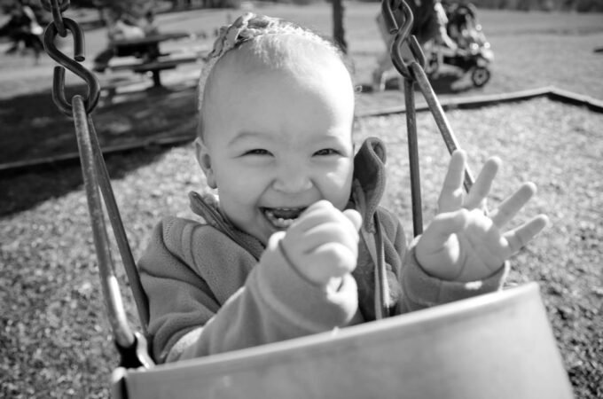 A happy child smiles and reaches from a playground bucket swing. Black and white close-up of metal chains and metal hooks attached to a swing.