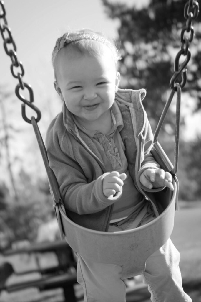 A smiling toddler in a bucket swing on an outdoor playground, captured in black and white. Details of the swing chains and the child's cozy jacket.