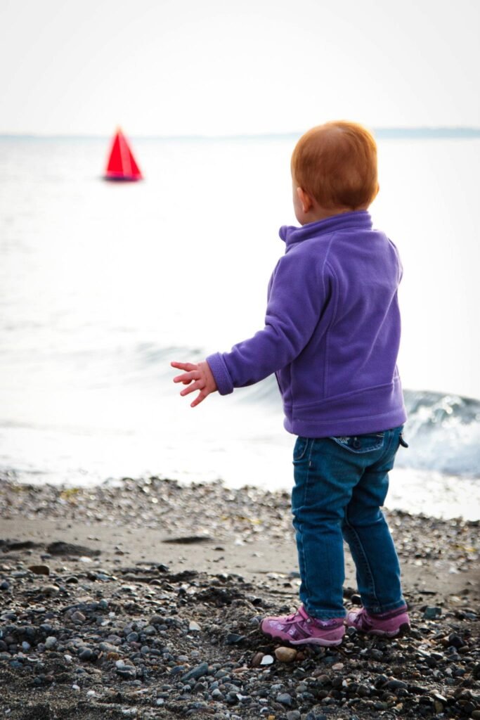 A child in a purple jacket stands on a rocky shore, looking towards a red marker floating in the water.