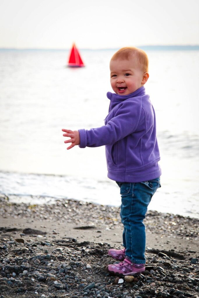 A smiling toddler in a purple jacket and pink boots plays on a pebble beach with calm water in the background.