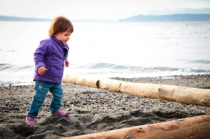 A toddler wearing a purple jacket and blue pants walks on a beach with driftwood logs and the sea in the background, capturing a moment of exploration.