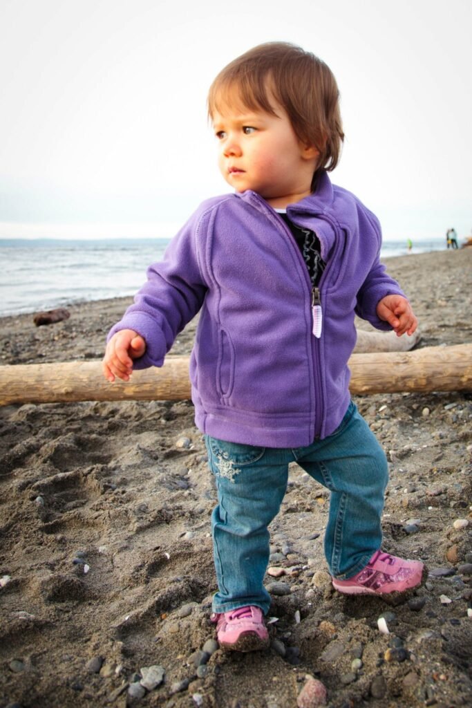 A little one in a purple fleece jacket and pink shoes takes wobbly steps on a Pacific Northwest beach, with a long driftwood log behind them.