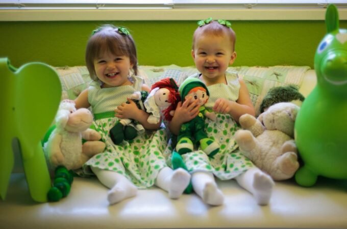 Two young children play with handmade dolls and stuffed animals in a green-themed room.