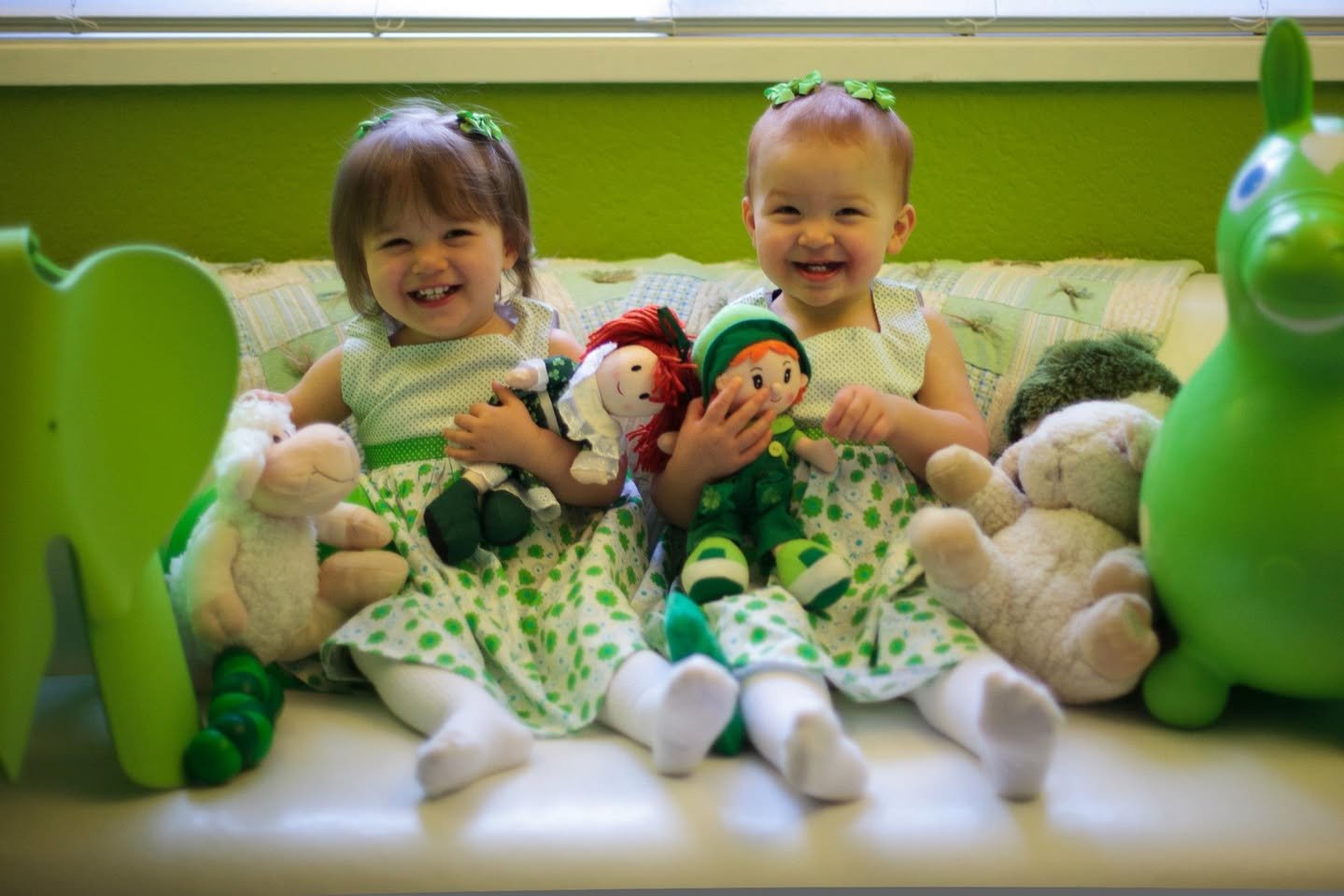 Two young children play with handmade dolls and stuffed animals in a green-themed room.