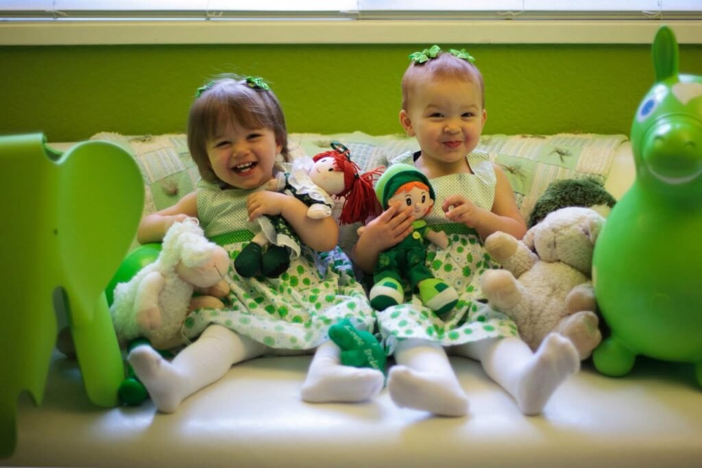 Two young children in green Christmas outfits smile surrounded by stuffed animals and holiday decorations indoors.