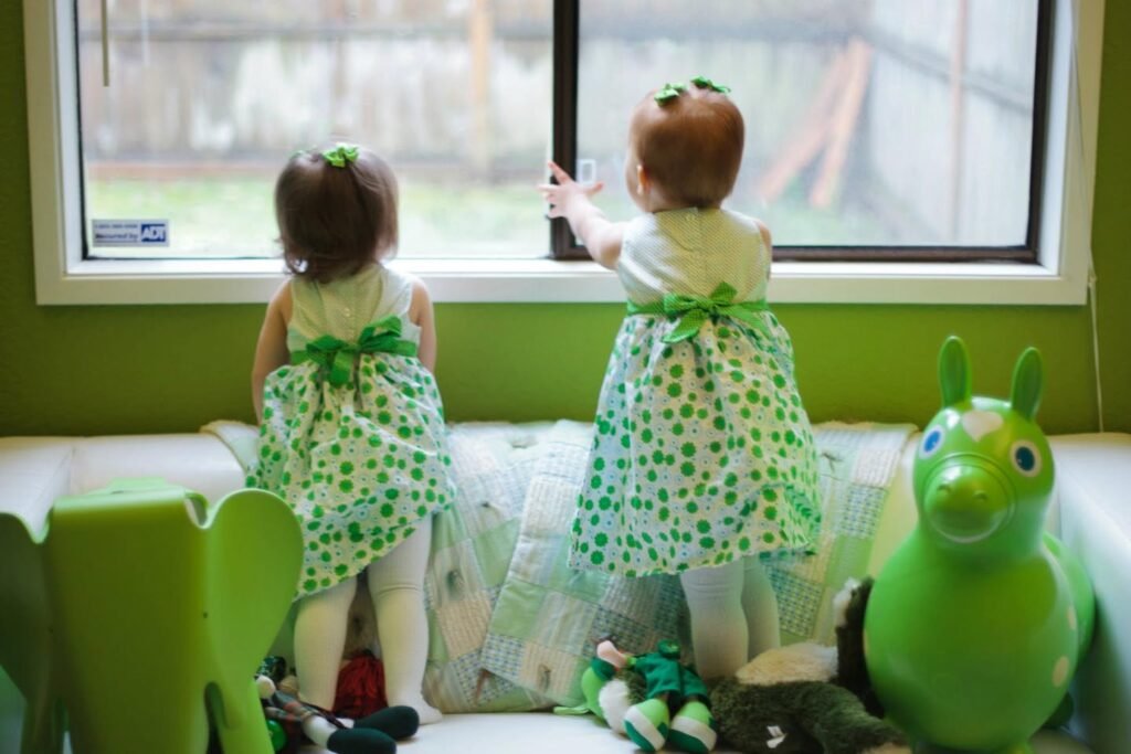Little ones in green polka dot dresses looking out a nursery window, next to a green toy