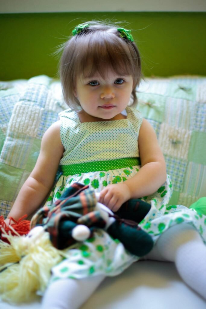 A toddler wearing a polka-dot dress and green hair accessories sits on a couch.