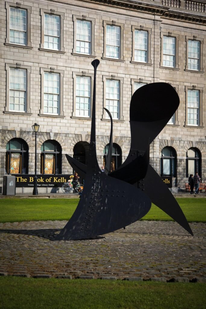 A close-up view of the ornate exterior windows of the Book of Kells museum in Trinity College Dublin, with a modern sculpture in the foreground.