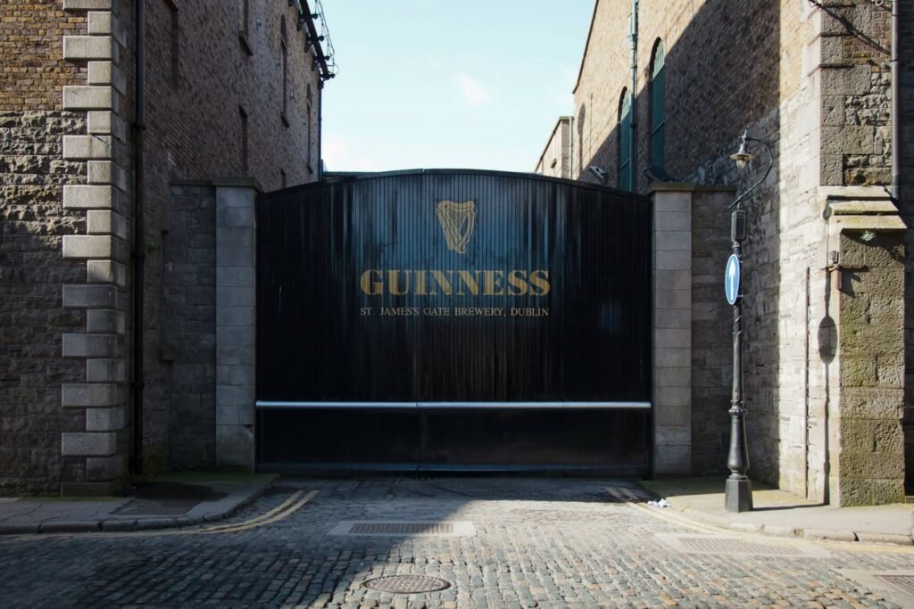 A street view of the iconic Guinness Storehouse entrance featuring the famous harp logo on a dark gate. The cobblestone path leads up to the historic landmark.