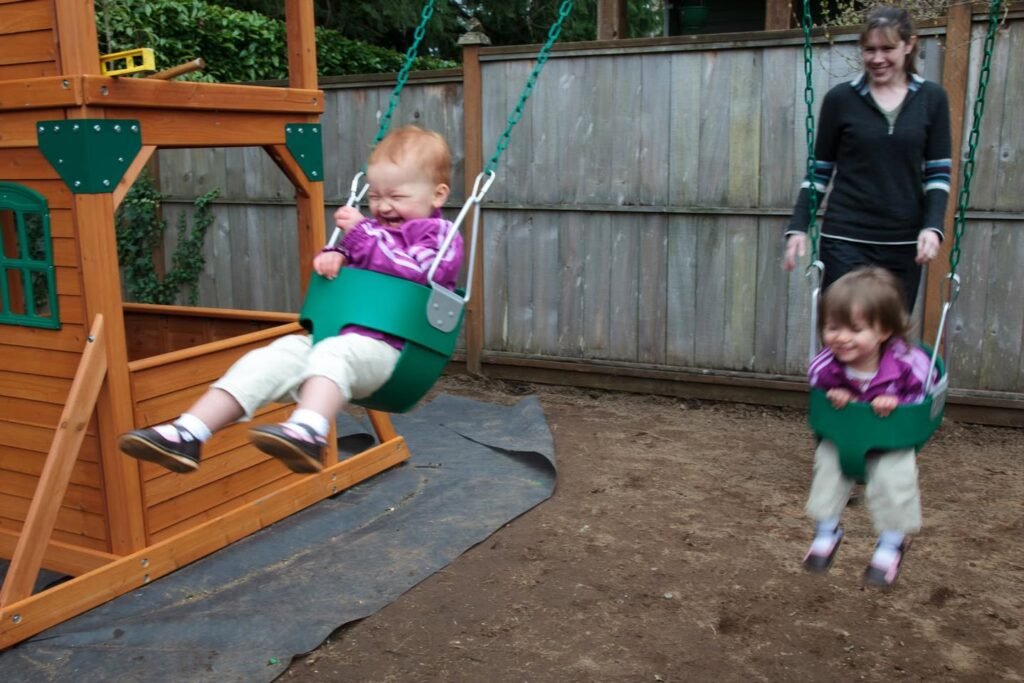 Young children enjoying green buckle swings on a wooden playset in a backyard, with a wooden fence and hedge in the background.