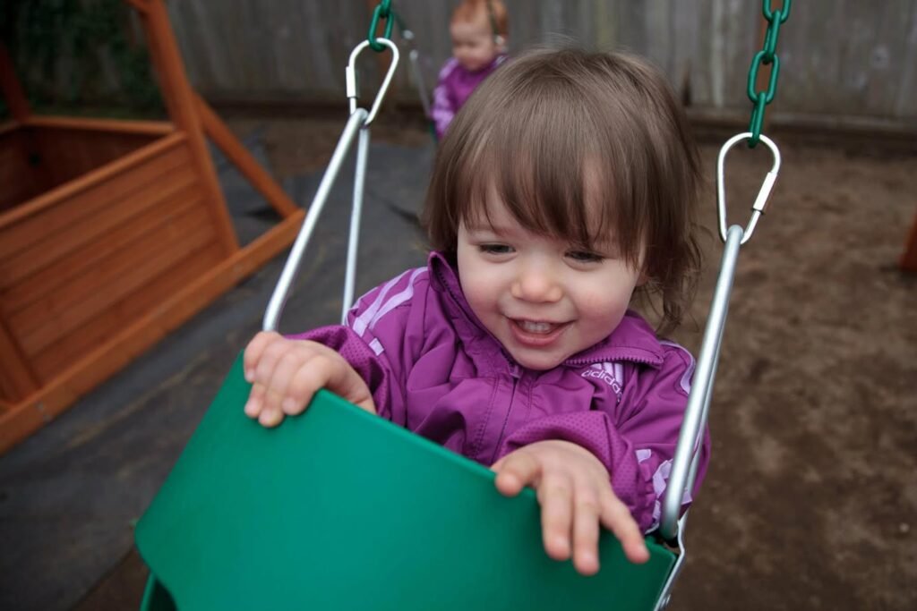A smiling toddler in a purple jacket holding on to a green swing outdoors.