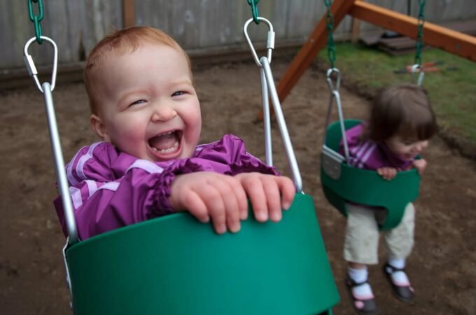 A baby smiles widely while riding a green swing in a backyard.