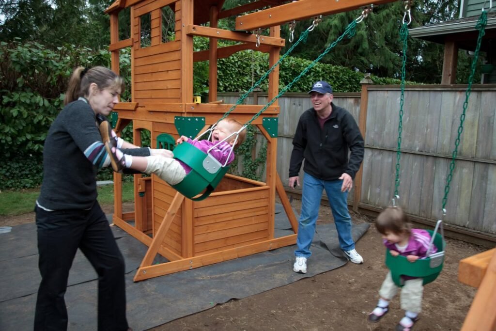 A woman pushes a child on a bucket swing in a backyard play set while another person watches nearby.