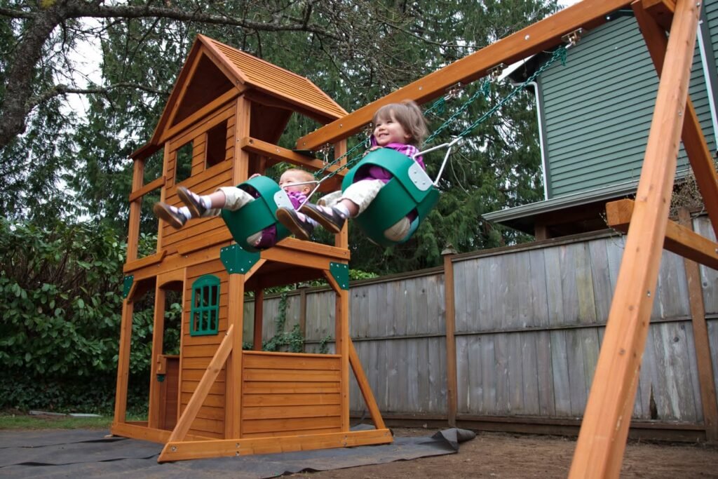 A child swings on a wooden play set in a backyard surrounded by trees and greenery.