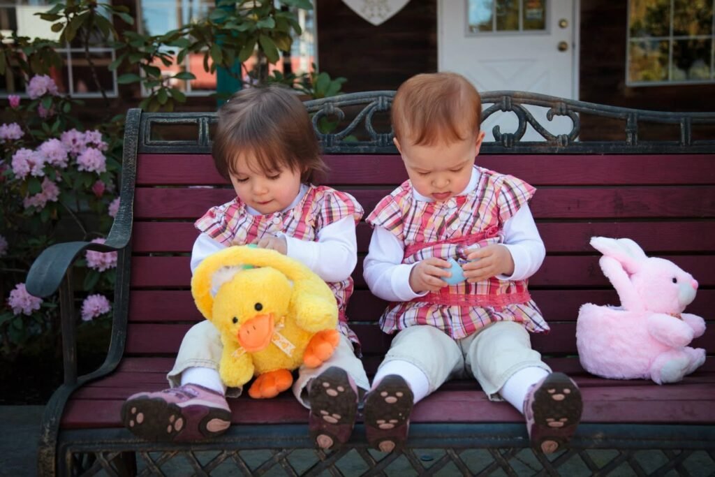 Young children sit on a porch bench holding Easter plush toys, a yellow duck and a pink bunny, surrounded by blooming pink flowers.