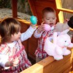 Two toddlers in matching outfits play with Easter eggs on a wooden play structure. A pink plush bunnies sits nearby.