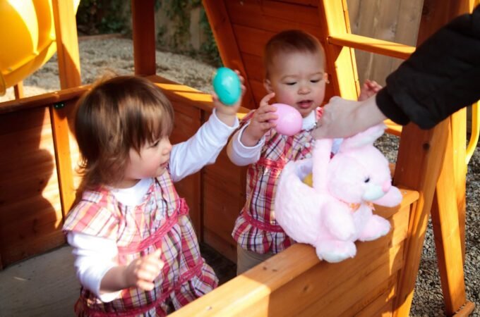Two toddlers in matching outfits play with Easter eggs on a wooden play structure. A pink plush bunnies sits nearby.