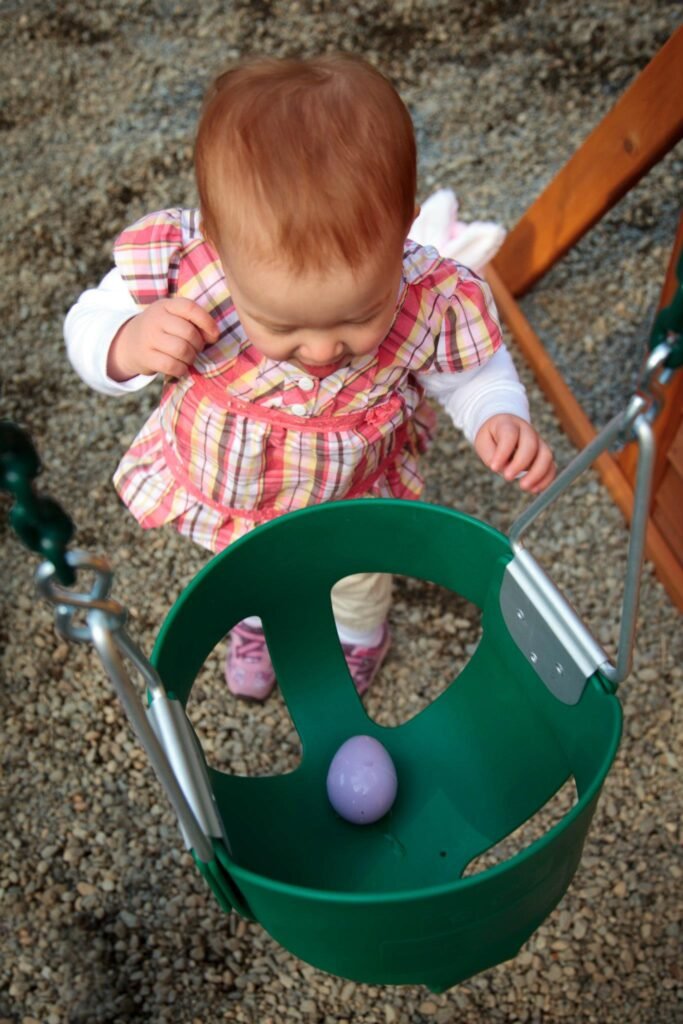 A baby wears a red plaid dress and looks down at a purple egg inside a green bucket. The outdoor gravel surface suggests an Easter egg hunt or backyard playtime.