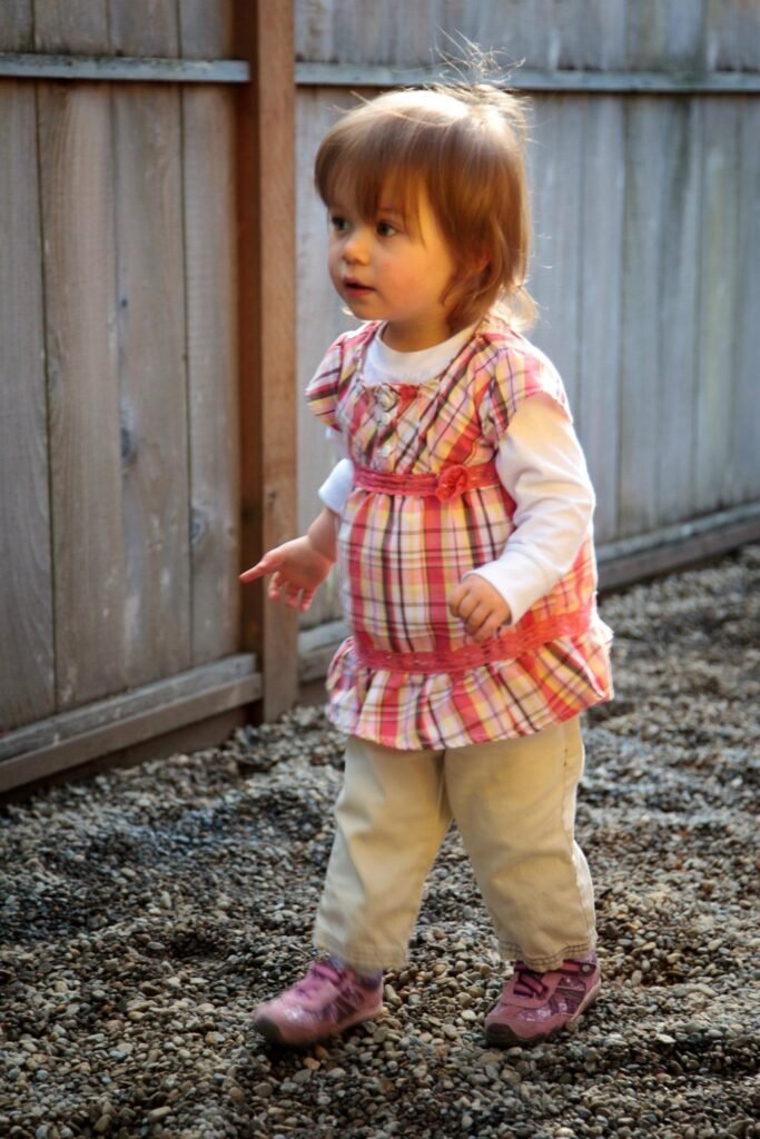 A child in a plaid dress explores the backyard by the wooden fence.