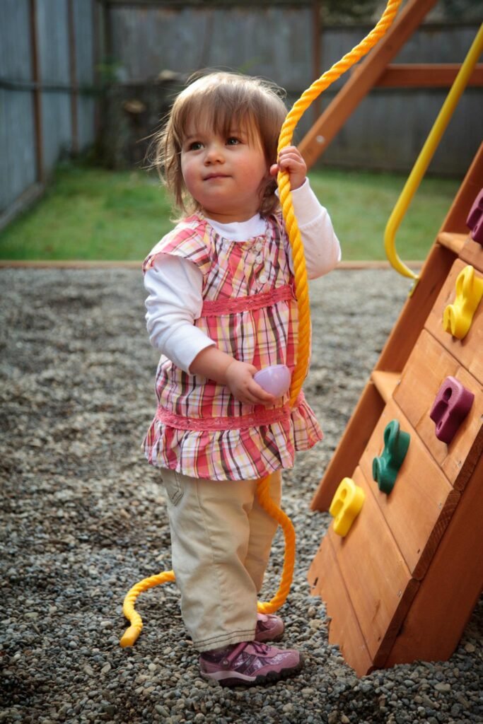 A toddler holding a yellow rope next to a wooden climbing gym. Outdoor playset with colorful holds on wood.