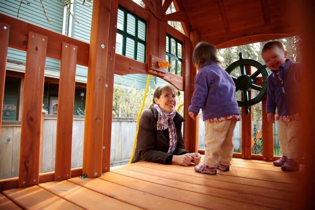 A parent sits on the wooden floor of a backyard play cabin while children play with a ship's wheel.