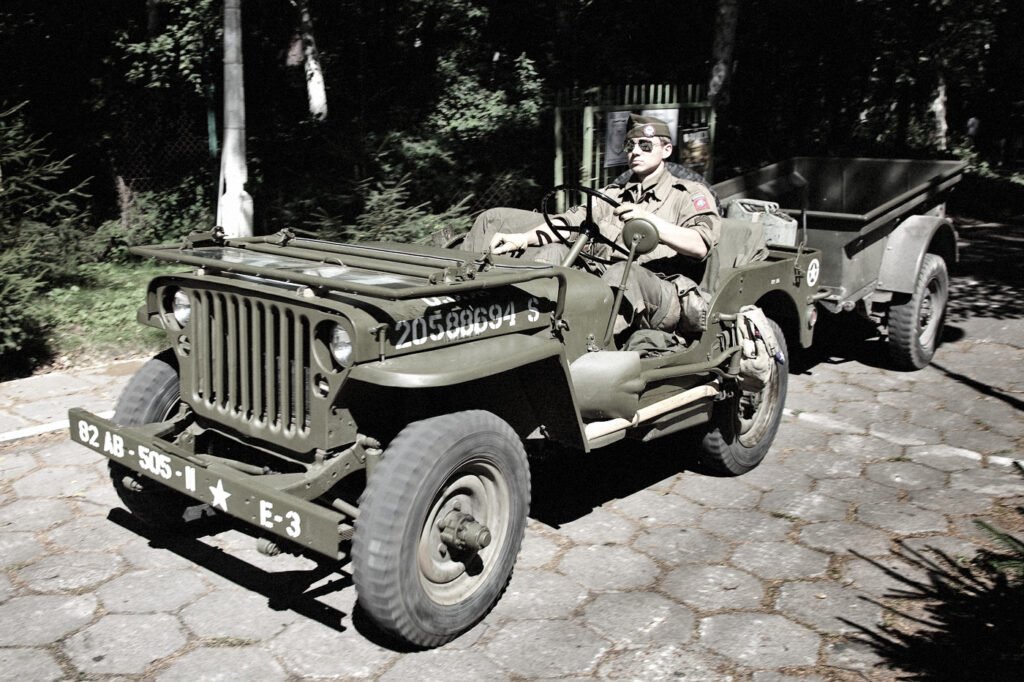 A vintage olive green Jeep, possibly from World War II, parked on cobblestones. A man in uniform sits in the driver's seat, holding the steering wheel.
