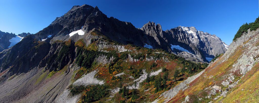 A stunning view of rugged mountains with snow-capped peaks and vibrant autumn foliage on the slopes. The clear blue sky complements the natural beauty of the landscape.