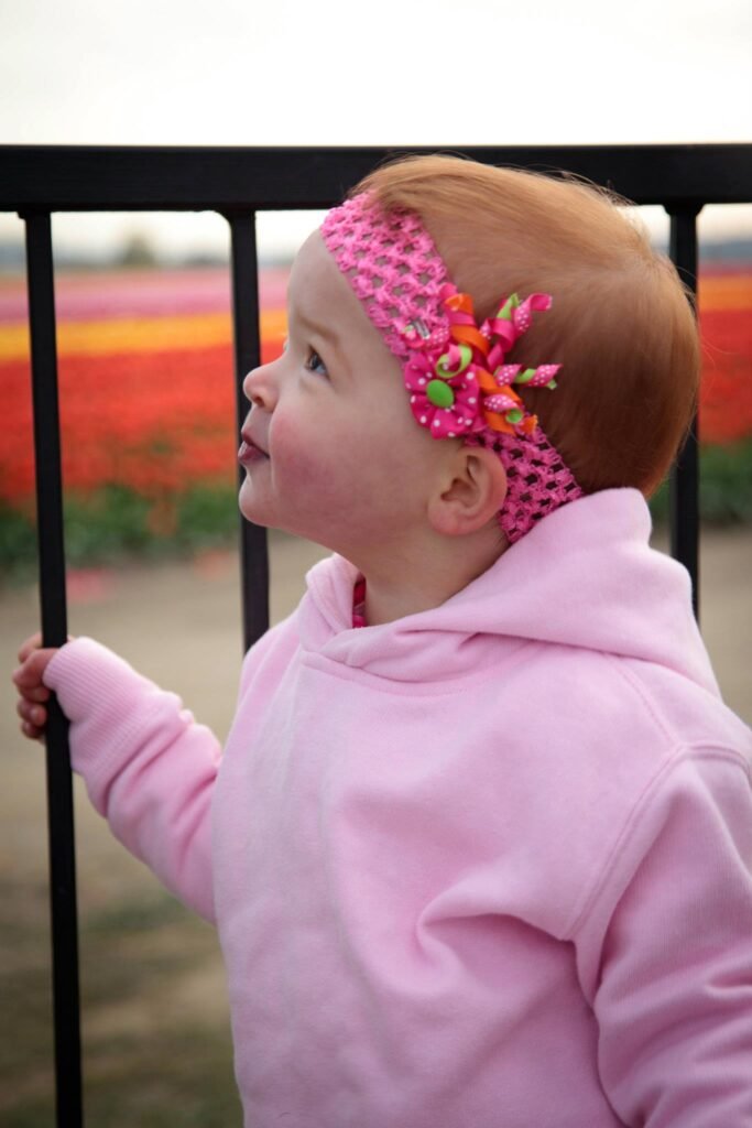 A young child in a pink fleece outfit and a knitted pink headband with flowers stands by a black railing. The background shows vibrant flower rows.