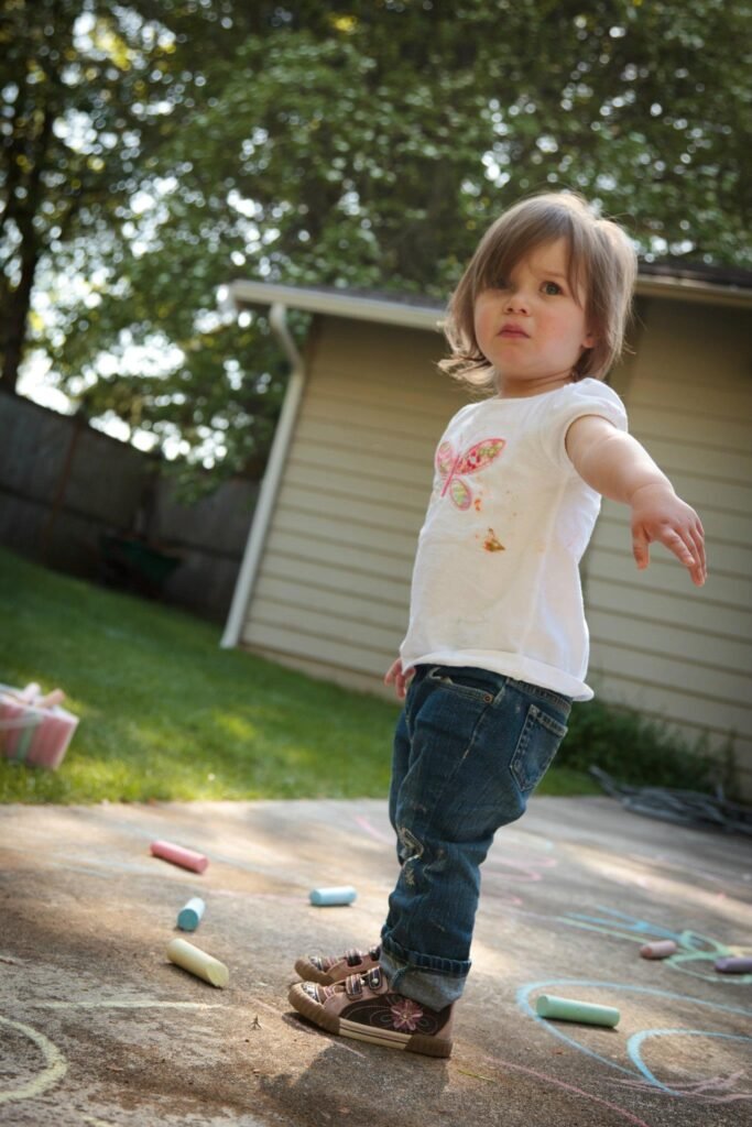 A toddler stands on a sunny patio surrounded by colorful wax crayons and chalk marksmarks. The young child has small hands outstretched as they explore their surroundings.