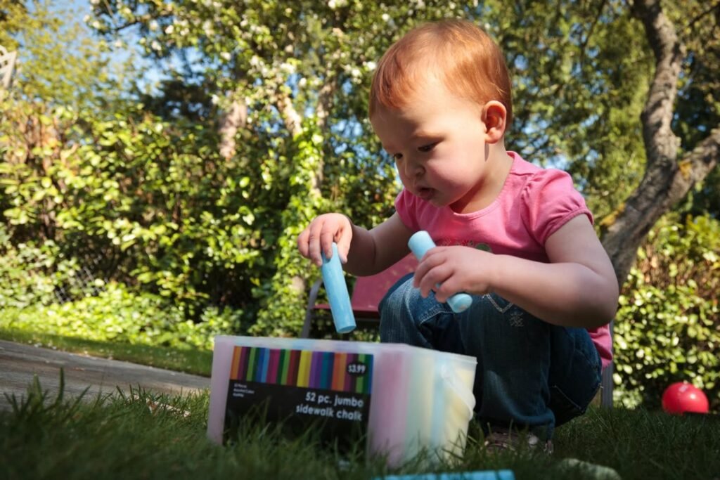 A toddler in a pink shirt crouches on the grass, holding blue chalk. The scene captures outdoor play on a sunny day with trees and a path in the background.