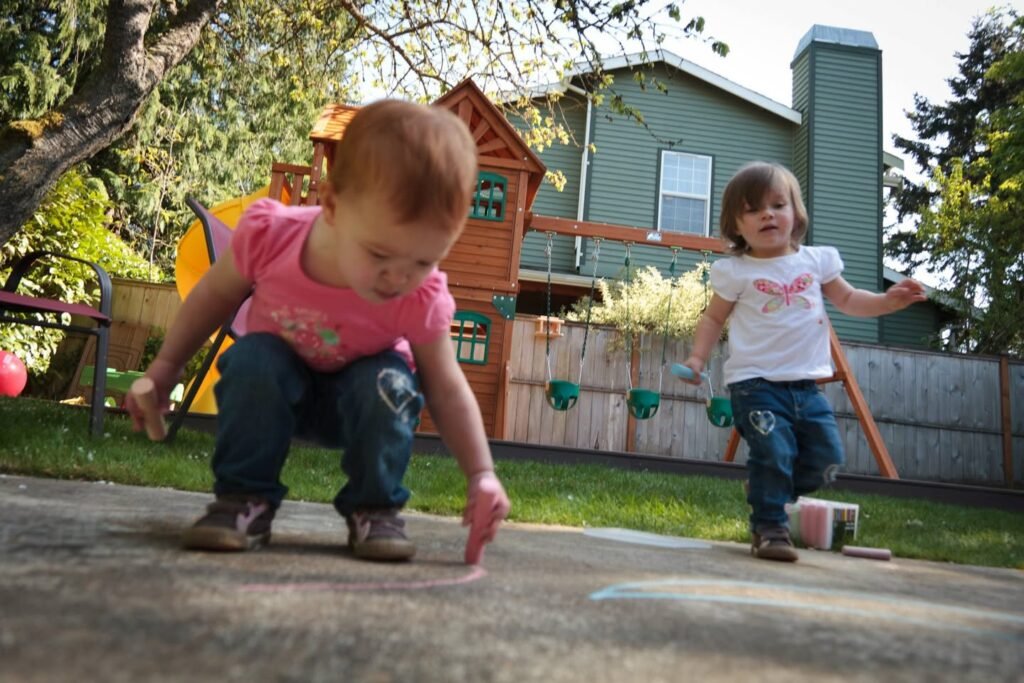 Two children playing with chalk on a driveway in a sunny backyard setting.