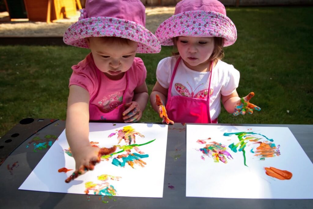 Two children in matching pink hats create colorful finger paintings on an outdoor picnic table.
