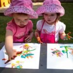 Two children in matching pink hats create colorful finger paintings on an outdoor picnic table.