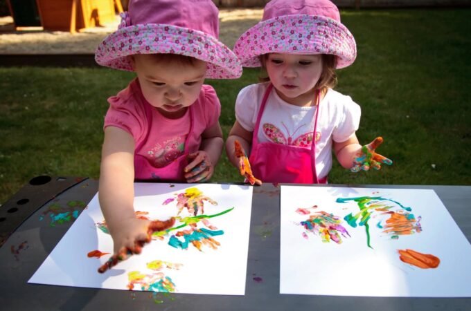 Two children in matching pink hats create colorful finger paintings on an outdoor picnic table.