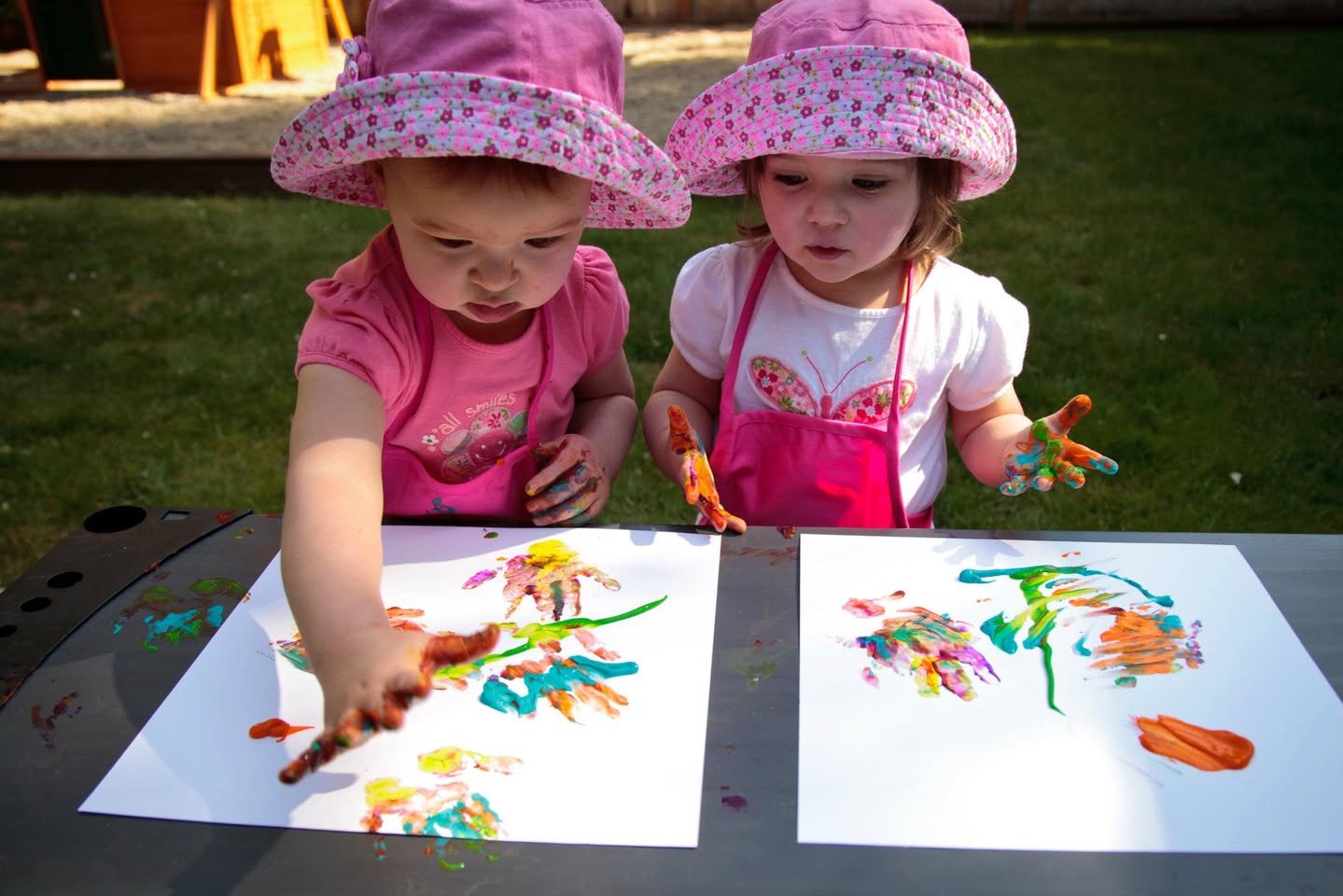 Two children in matching pink hats create colorful finger paintings on an outdoor picnic table.