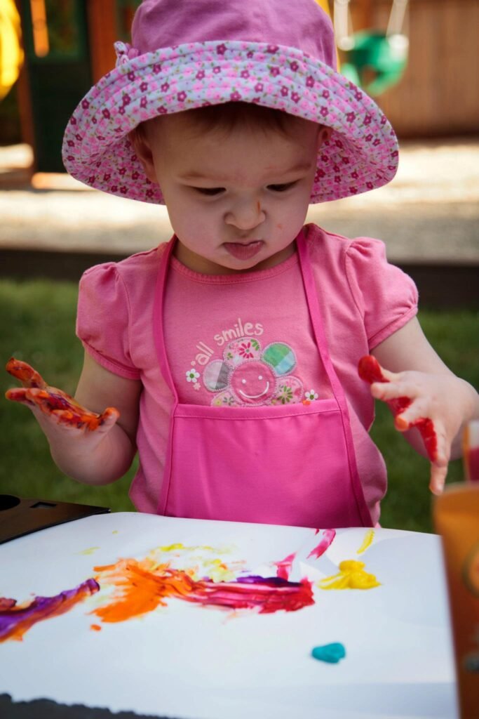 Toddler in a pink hat and outfit painting with colorful smears of paint on a white board outdoors.