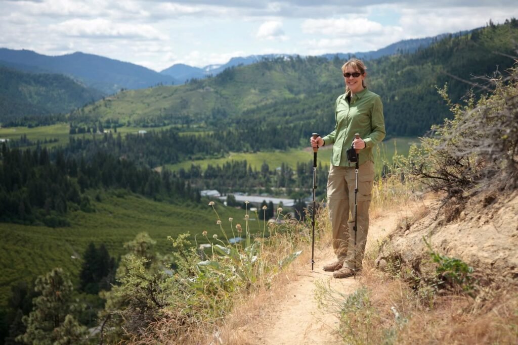 A person is hiking on a dirt trail overlooking a mountain valley filled with trees and distant buildings.