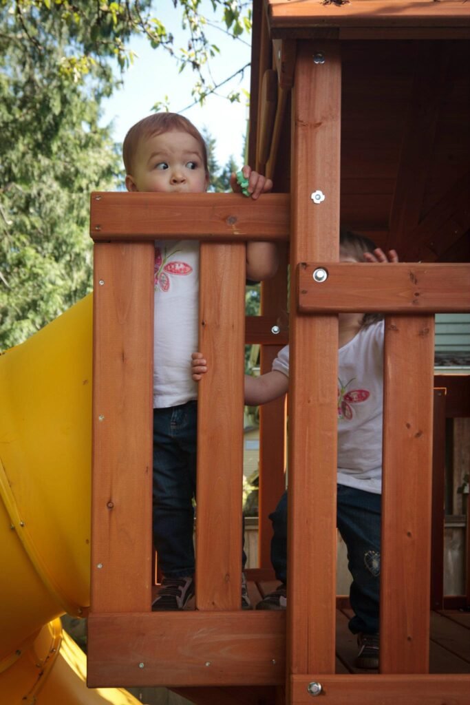 Children playing inside a wooden playhouse with a yellow slide, surrounded by greenery.