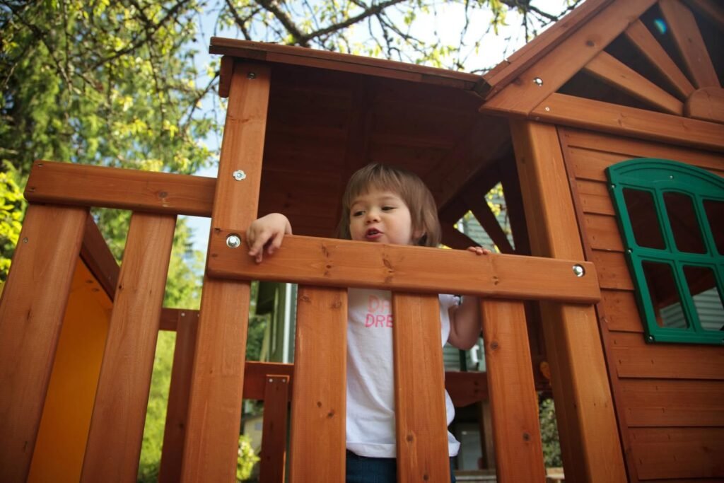 A child peeks out from the railing of a wooden playhouse surrounded by lush trees.