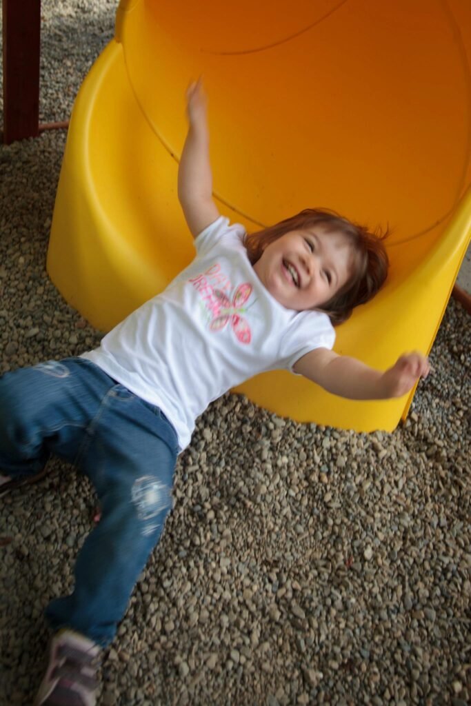 A joyful child is lying at the base of a bright yellow plastic slide on a gravel surface at a playground. The scene captures a playful and energetic moment.