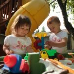Children enjoy a plastic play kitchen set outdoors under a yellow slide in a sunny backyard with trees and a wooden deck structure.