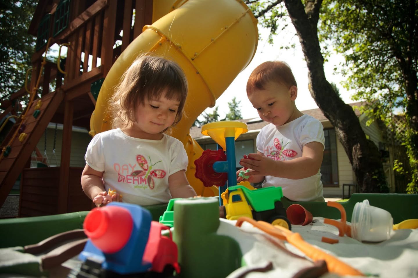Children enjoy a plastic play kitchen set outdoors under a yellow slide in a sunny backyard with trees and a wooden deck structure.