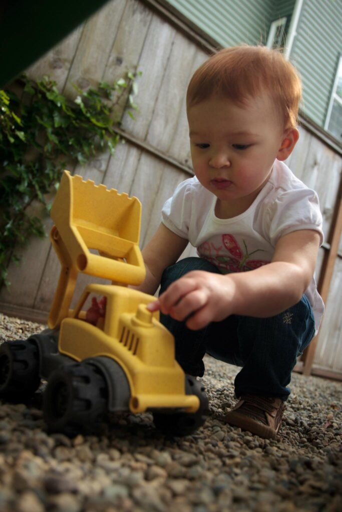 A toddler in a white shirt and blue pants crouchs near a yellow toy dump truck on gravel.