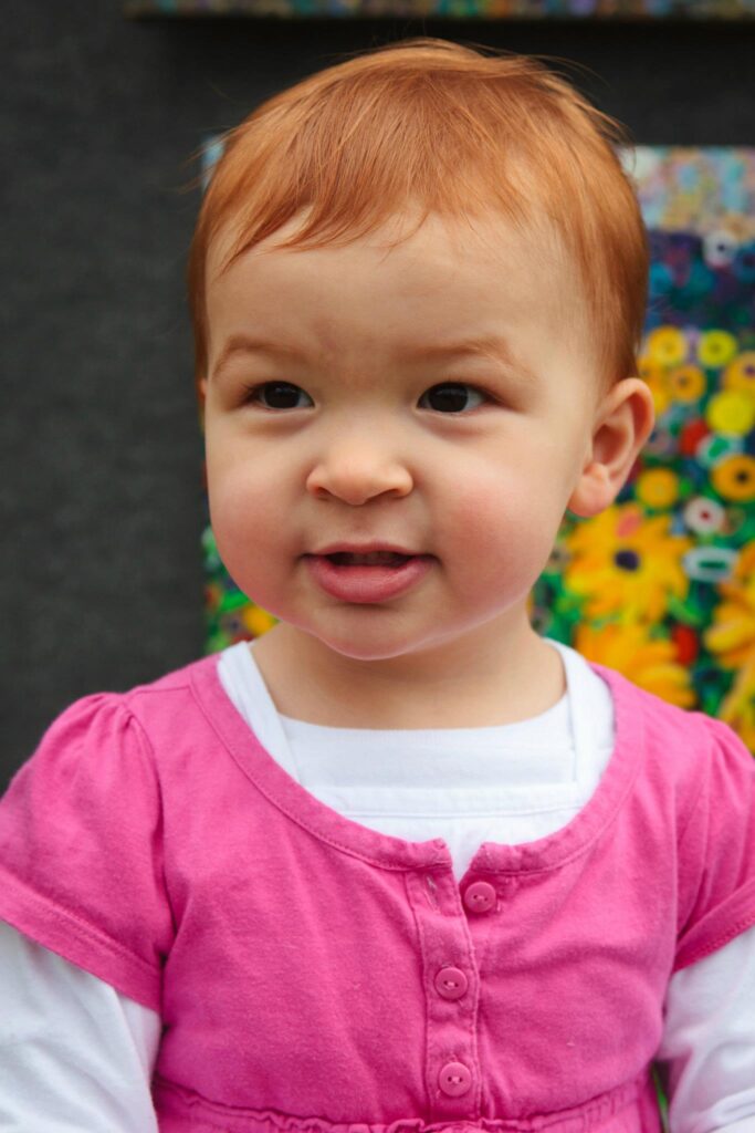 A young child with bright red hair wearing a pink blouse with white sleeves stands in front of a colorful floral mural.