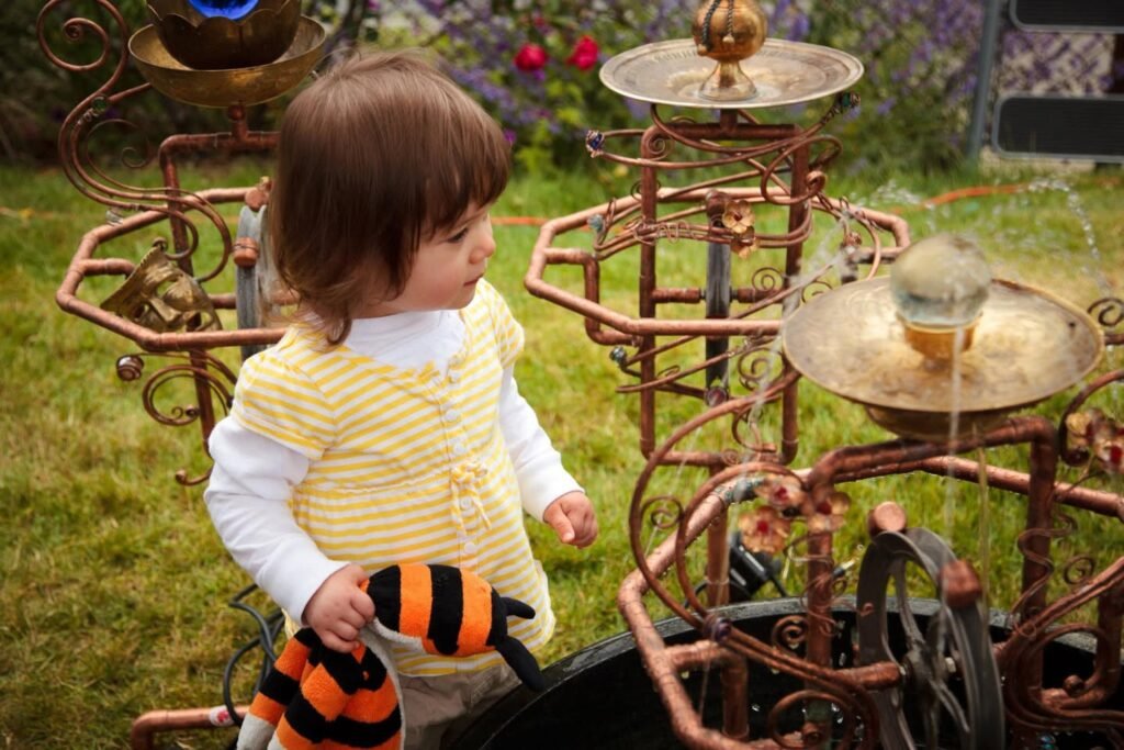 A child in a yellow cardigan examines ornate metal bird baths while holding a bumblebee plushie. Curled metal details and water features are shown in a backyard setting.