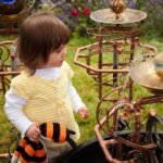 A child in a yellow cardigan examines ornate metal bird baths while holding a bumblebee plushie. Curled metal details and water features are shown in a backyard setting.